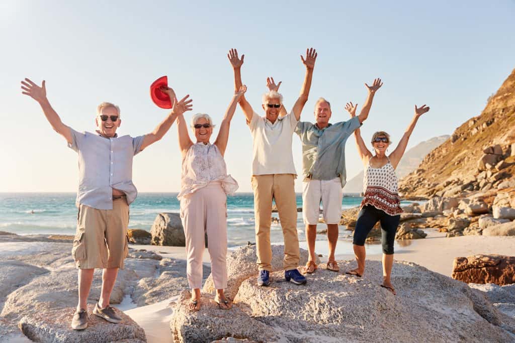 Happy seniors celebrating on the beach with arms raised and smiling under clear sky.